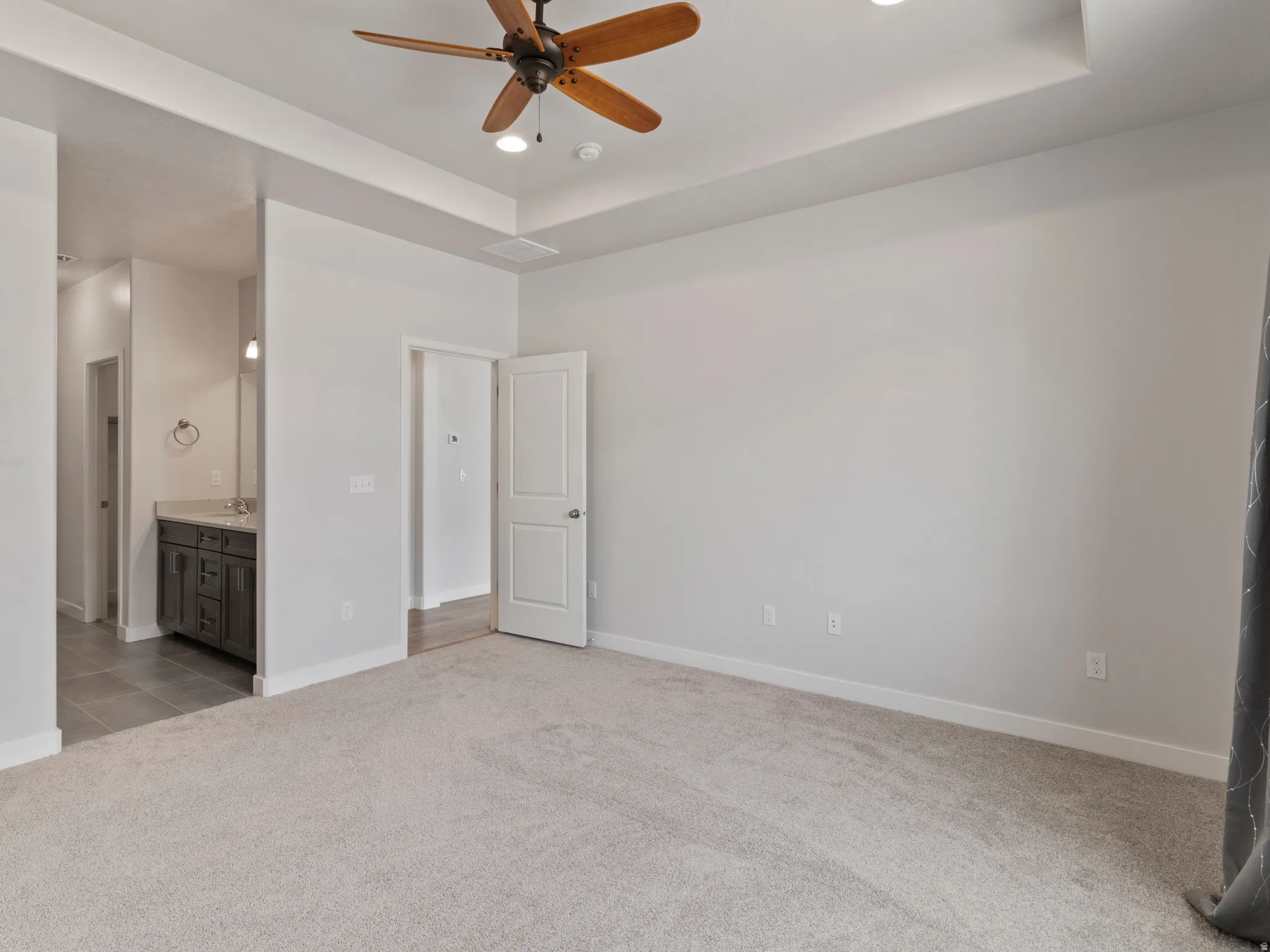 Tray ceiling with recessed lighting and a ceiling fan, plush carpeting, and an adjoining bathroom featuring a dark wood-finish vanity with a light-colored countertop