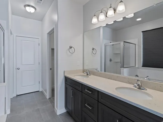 Dual sink vanity with stone countertop, tile flooring, and a glass-enclosed shower