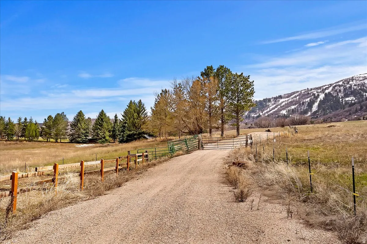 Gravel drive with a metal gate, leading to a sprawling property featuring mature trees and a distant mountain backdrop