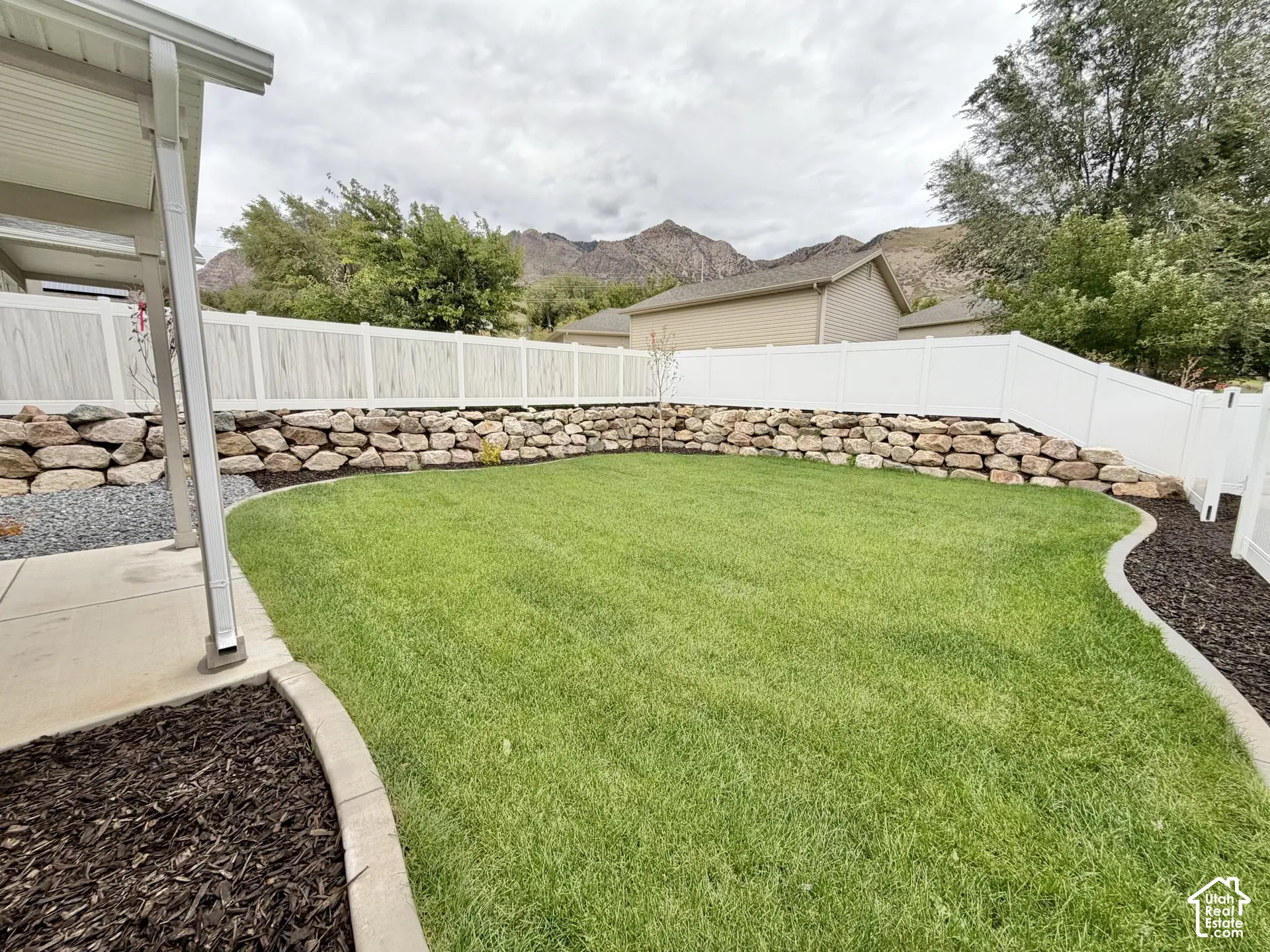 Backyard featuring a manicured lawn, stone retaining wall, white vinyl fencing, concrete patio, and mountain backdrop