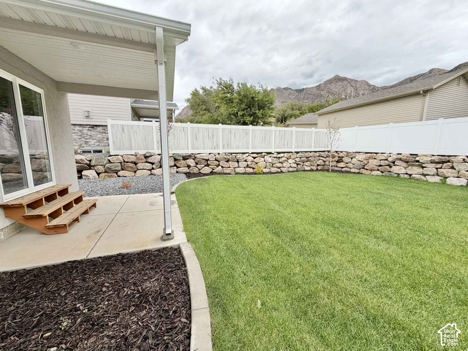 Covered concrete patio with wooden stairs, manicured lawn, stone retaining wall, white privacy fencing, and mountain backdrop