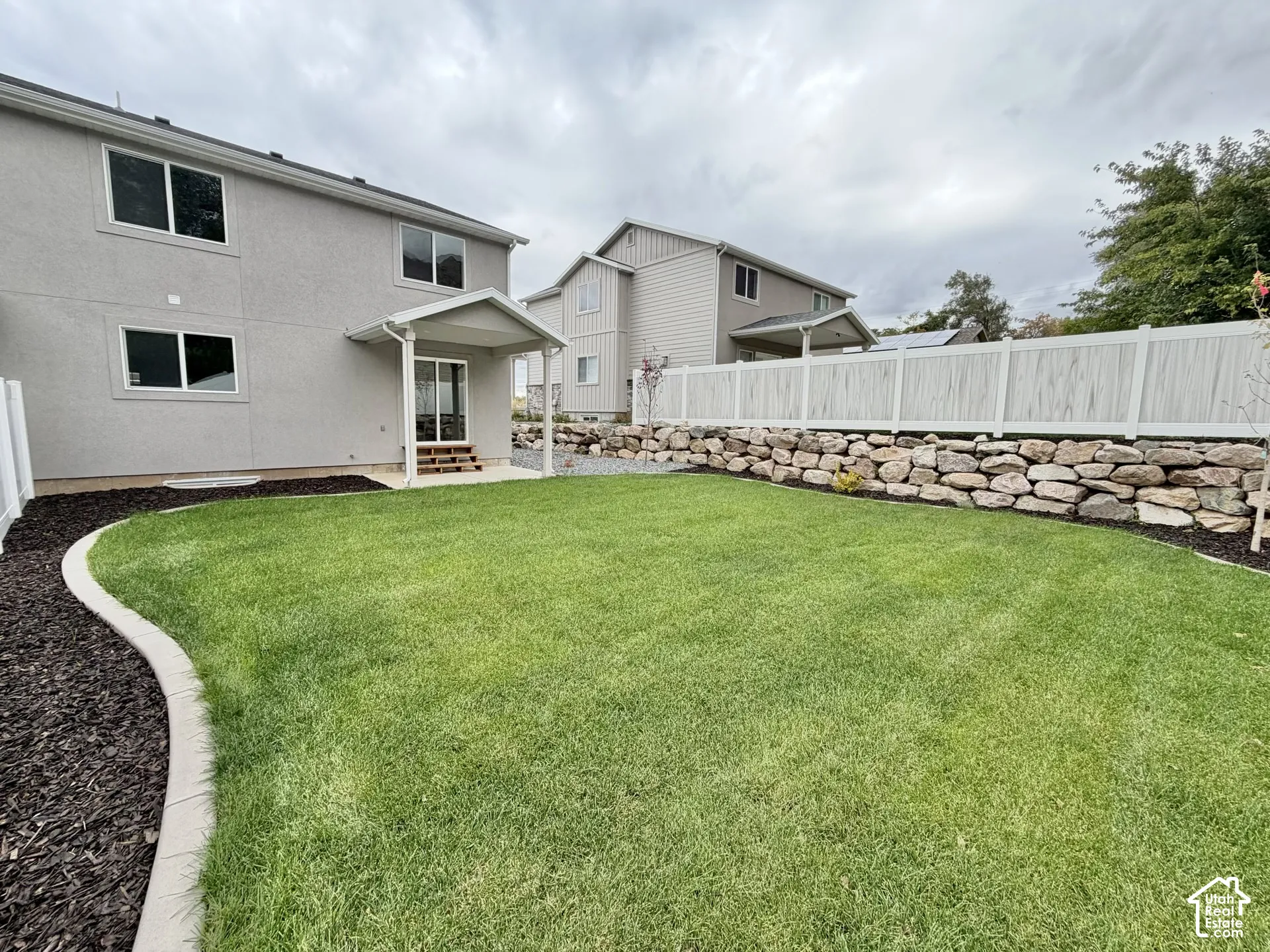 Backyard featuring a manicured lawn, stone retaining wall, concrete landscape edging, covered patio, and privacy fencing