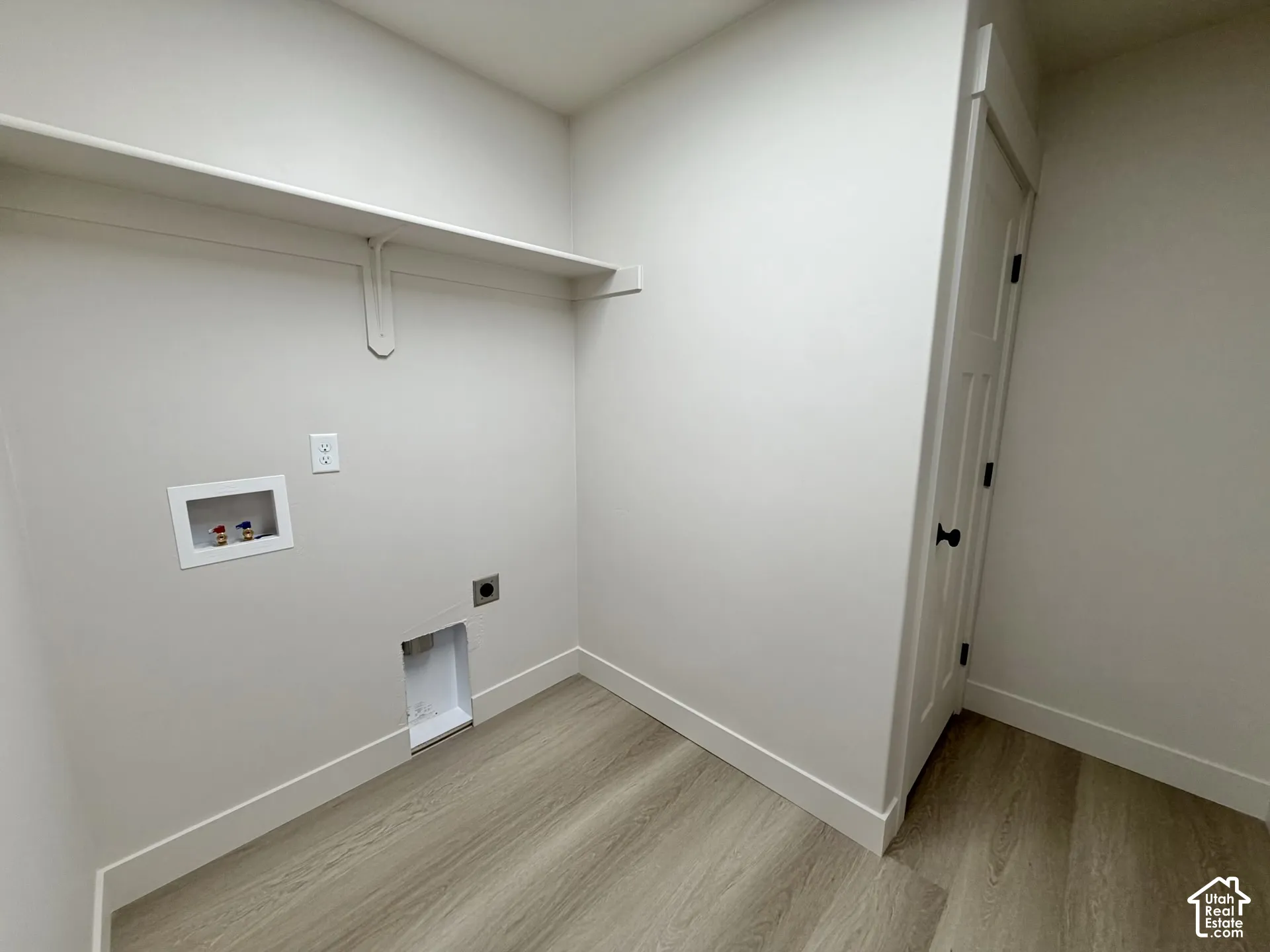 Utility area featuring wood-finish flooring, a white-paneled door with matte black hardware, and an overhead storage shelf