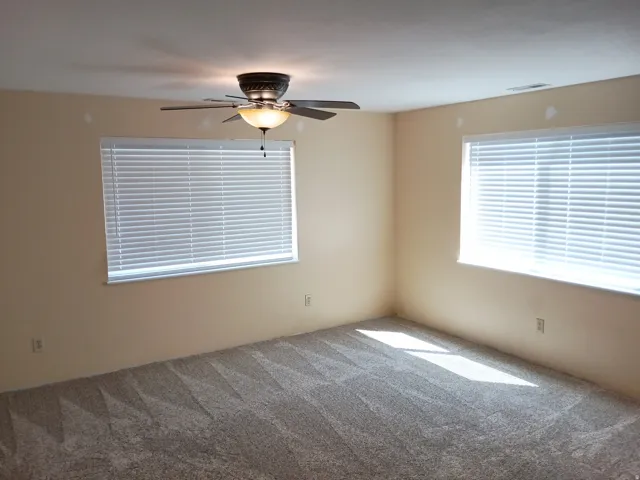 Neutral tone wall paint, two windows with blinds, ceiling fan with integrated lighting, and patterned carpet flooring