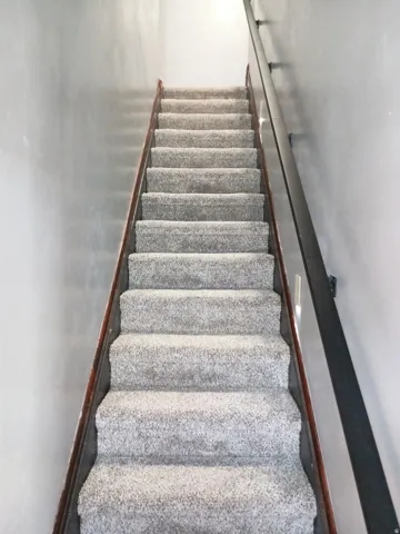 Staircase featuring light gray carpeted treads and risers, a stained wood stringer, and a black metal handrail