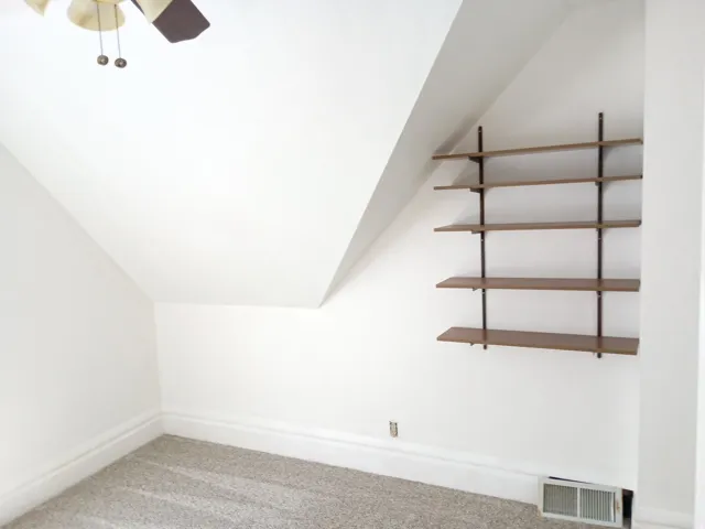 Sloped ceiling room featuring a ceiling fan, wall-mounted shelving, and carpeted flooring