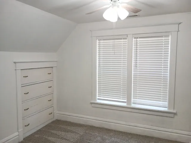 Ceiling fan with light fixture, built-in drawer storage, dual windows with blinds, and sloped ceiling