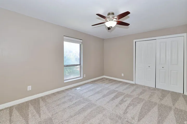 Carpeted flooring, neutral wall tones, window with roller shade, ceiling fan, and bi-fold closet doors