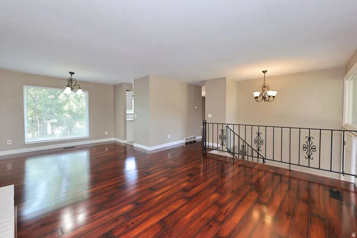 Rich wood-finish flooring, two chandeliers, expansive window with blinds, wrought iron railing, and neutral wall tones