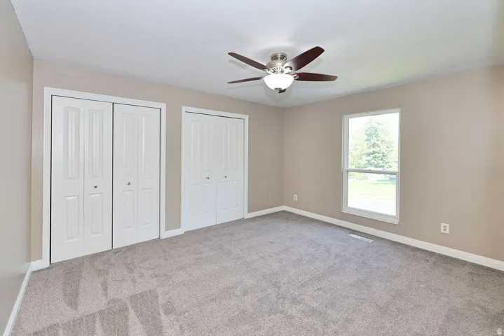 Neutral tone wall paint, light gray carpeting, ceiling fan with integrated lighting, multiple bi-fold closet doors, and a double-hung window