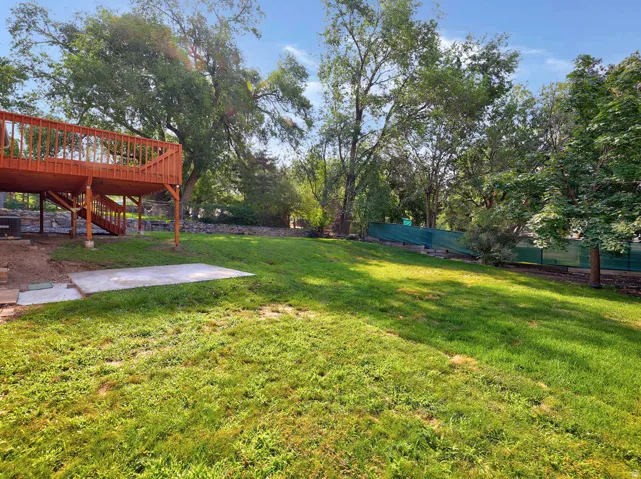 Expansive grassy yard with mature trees, featuring a raised wood deck with staircase access, and a concrete patio pad