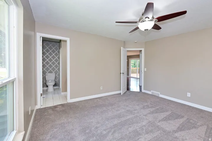 Neutral tone walls, grey patterned carpet, ceiling fan with light fixture, white trim, and a window with white casing