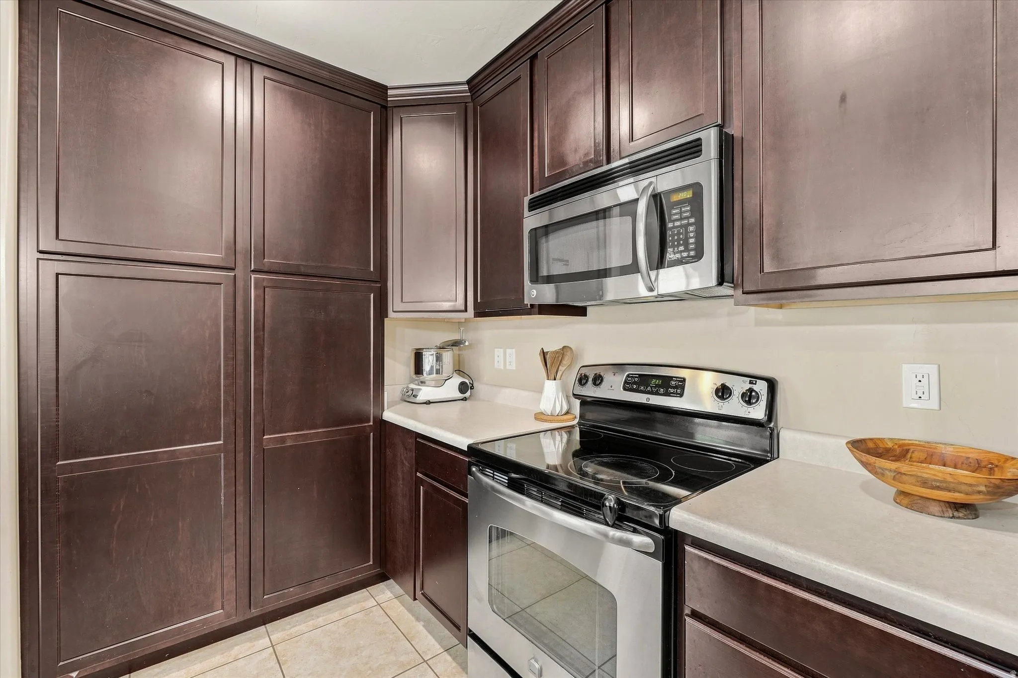 Dark wood-finish cabinetry, stainless steel microwave, electric range, light-toned countertops, and tiled flooring