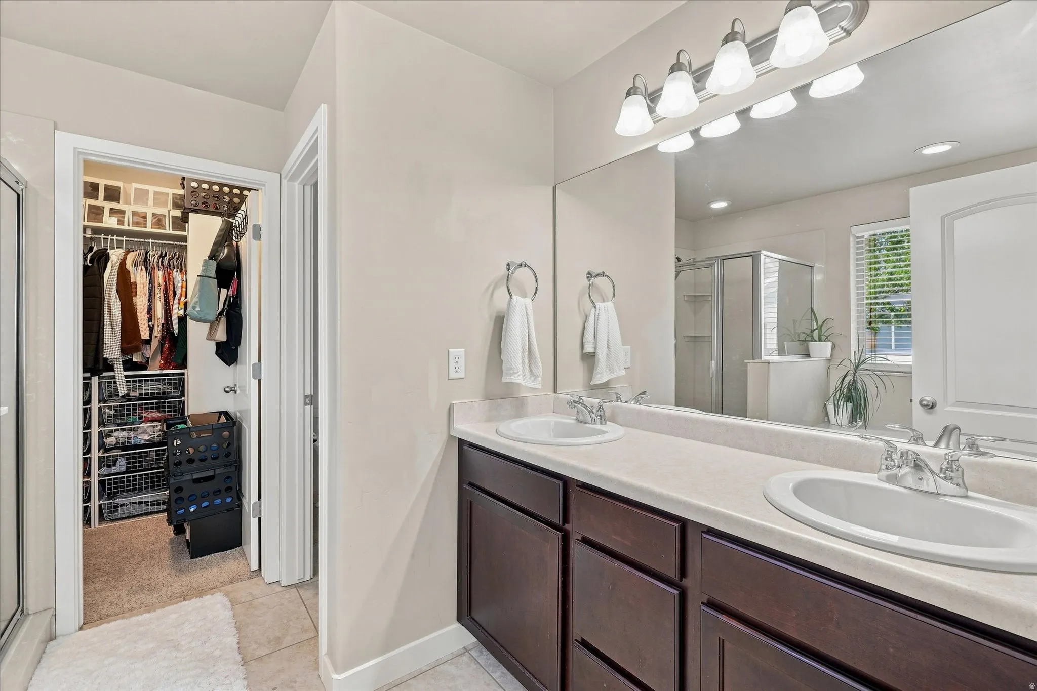 Dual vanity with integrated sinks, expansive wall mirror, dark wood-finish cabinetry, and a walk-in closet with built-in shelving