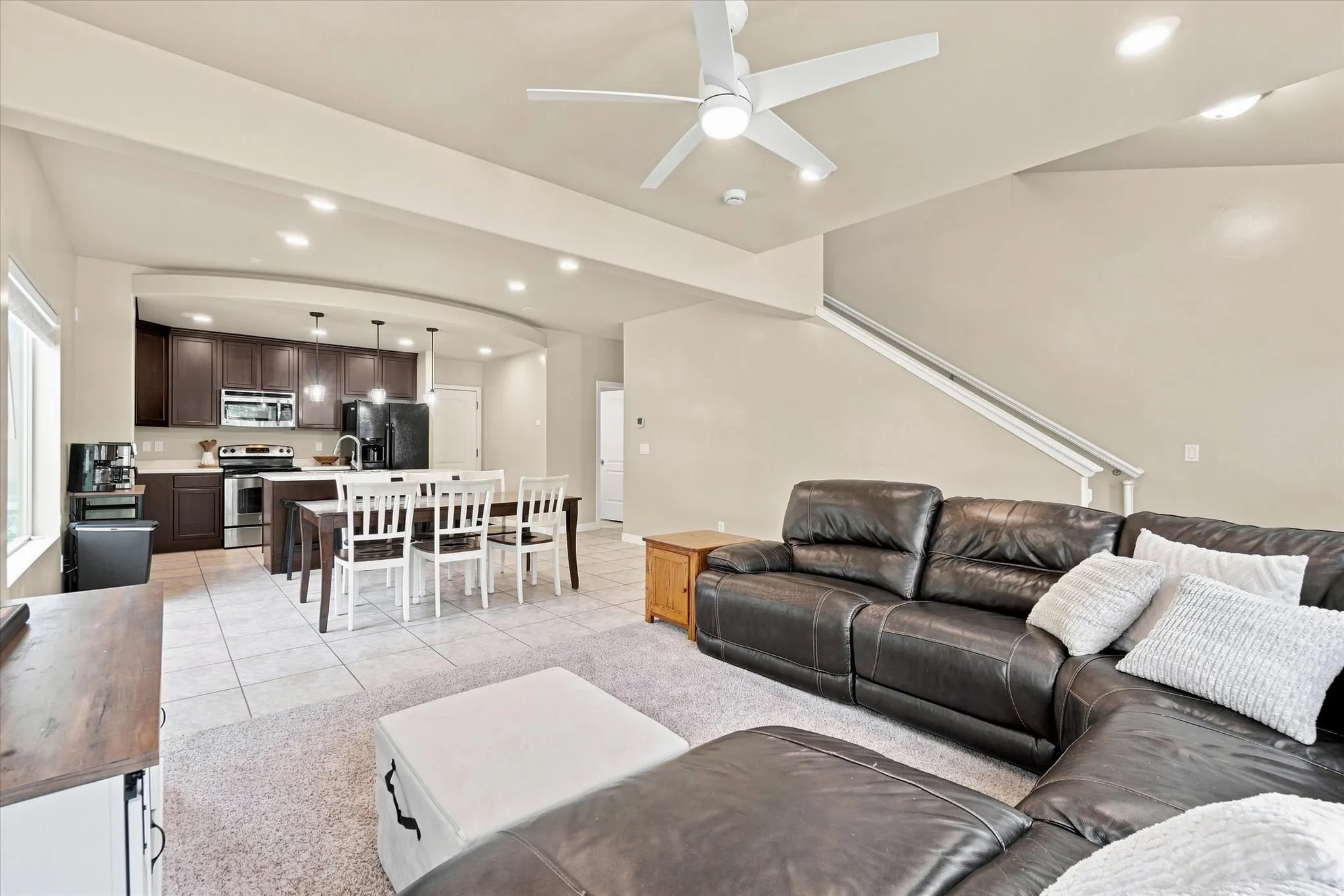 Open-concept living area with integrated kitchen and dining, featuring recessed lighting, a modern ceiling fan, and staircase access
