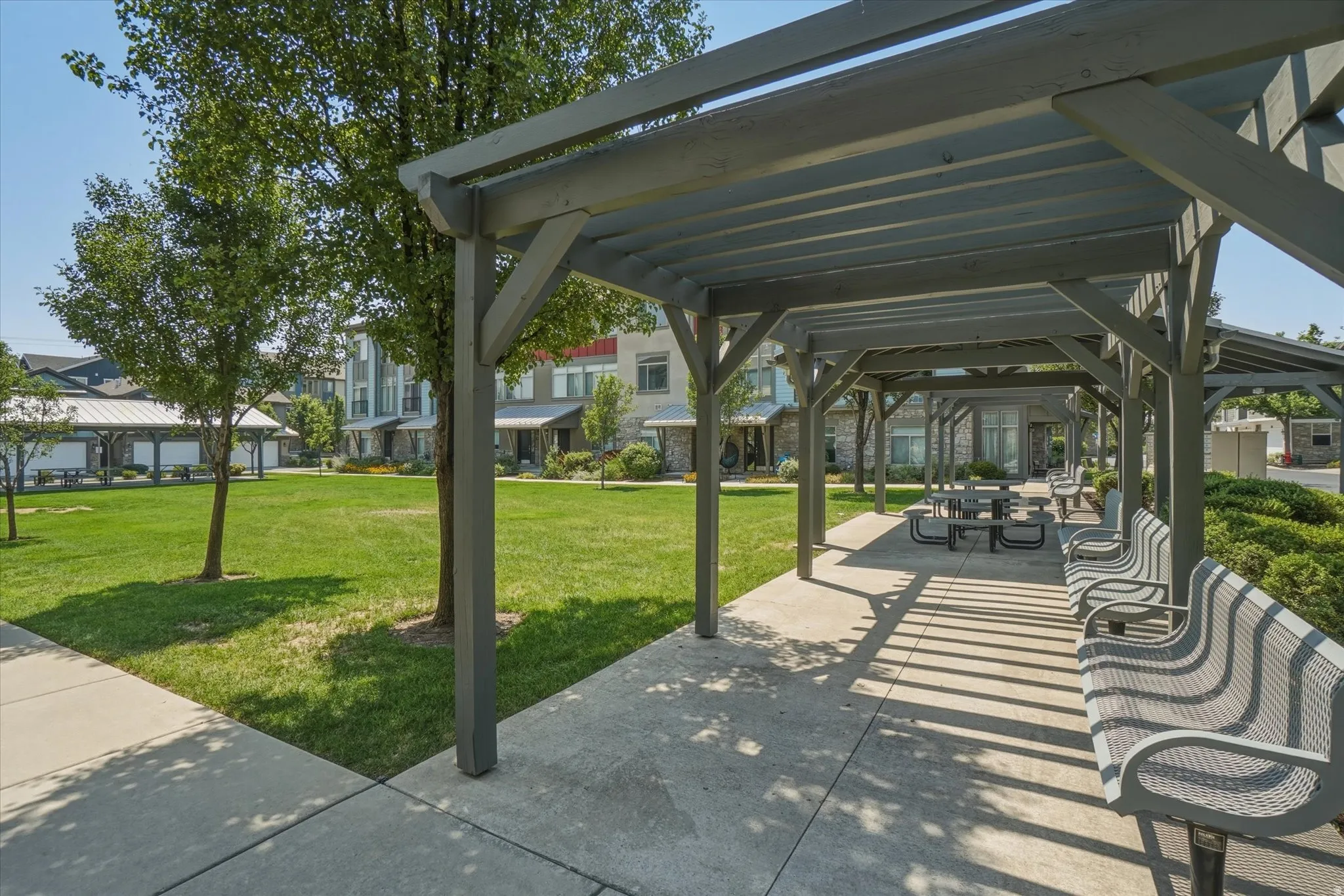 Covered outdoor seating area with multiple benches and picnic tables, surrounded by a manicured lawn and mature trees