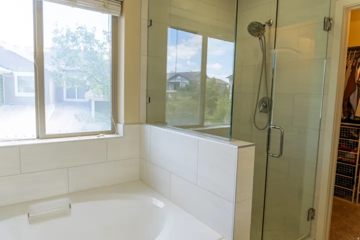Built-in bathtub, tiled surround, window with blinds, glass-enclosed shower, and a chrome showerhead