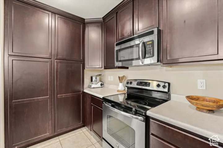 Dark wood-finish cabinetry, stainless steel microwave, electric range, light-toned countertops, and tiled flooring