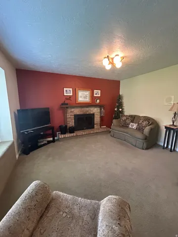 Carpeted living area featuring a brick-surround fireplace with a wood mantle, a textured ceiling, and a multi-bulb ceiling fixture