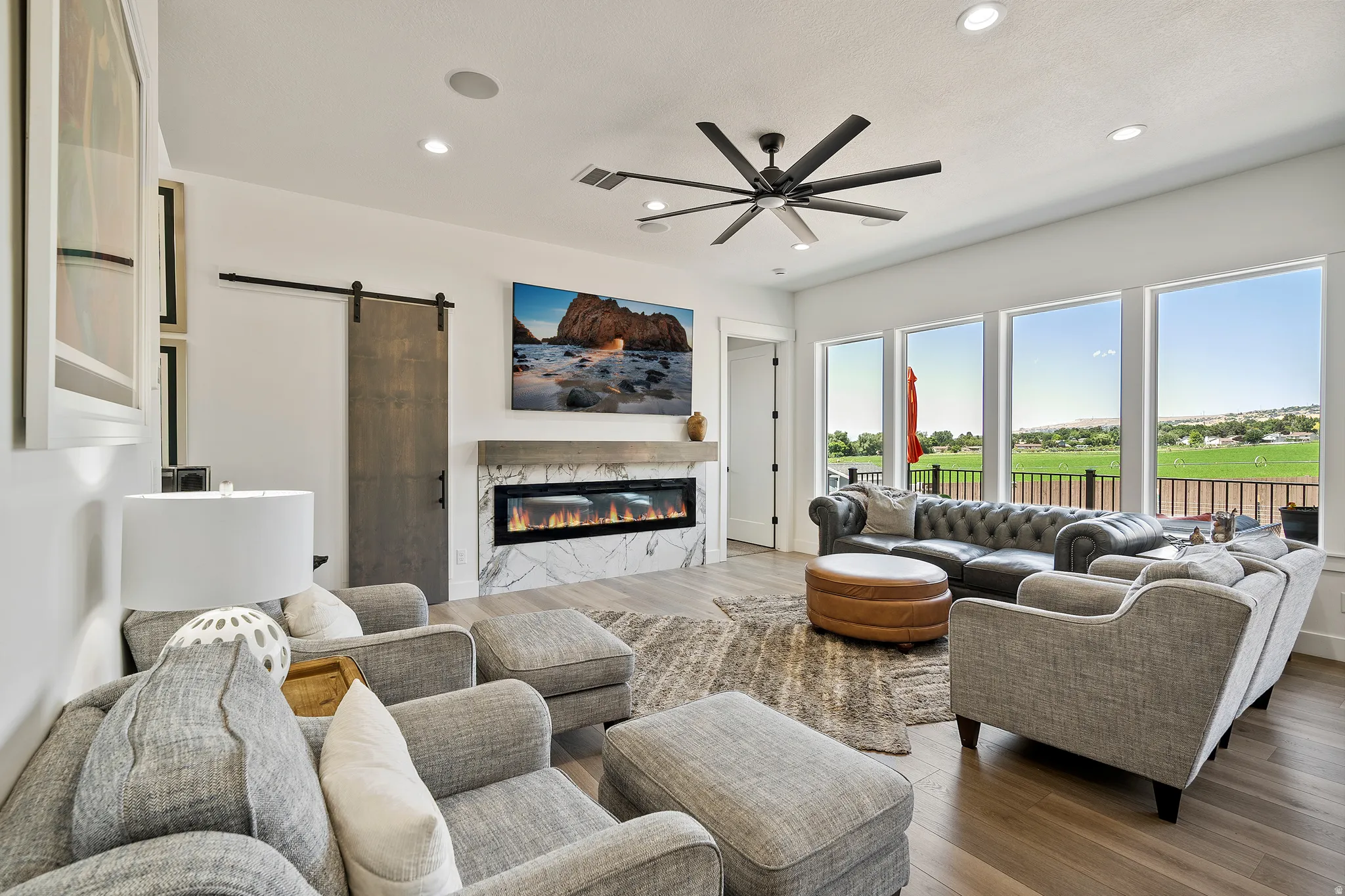 Spacious living area featuring wood-finish flooring, a linear fireplace with a stone surround, and abundant natural light from multiple large windows