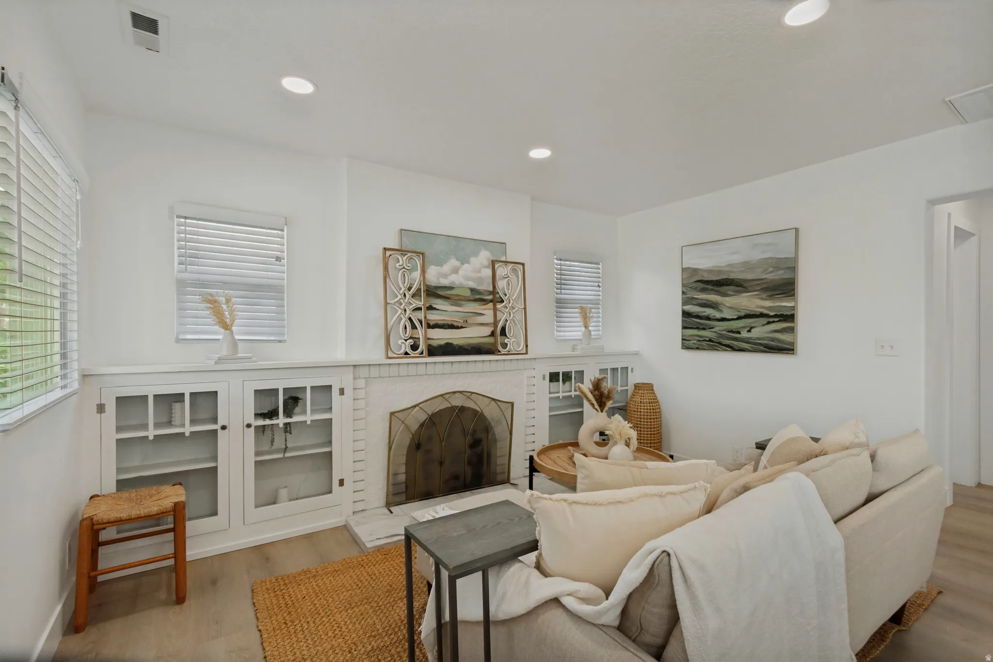 Living area featuring a white brick fireplace, built-in cabinetry with glass panel doors, recessed lighting, and wood-finish flooring