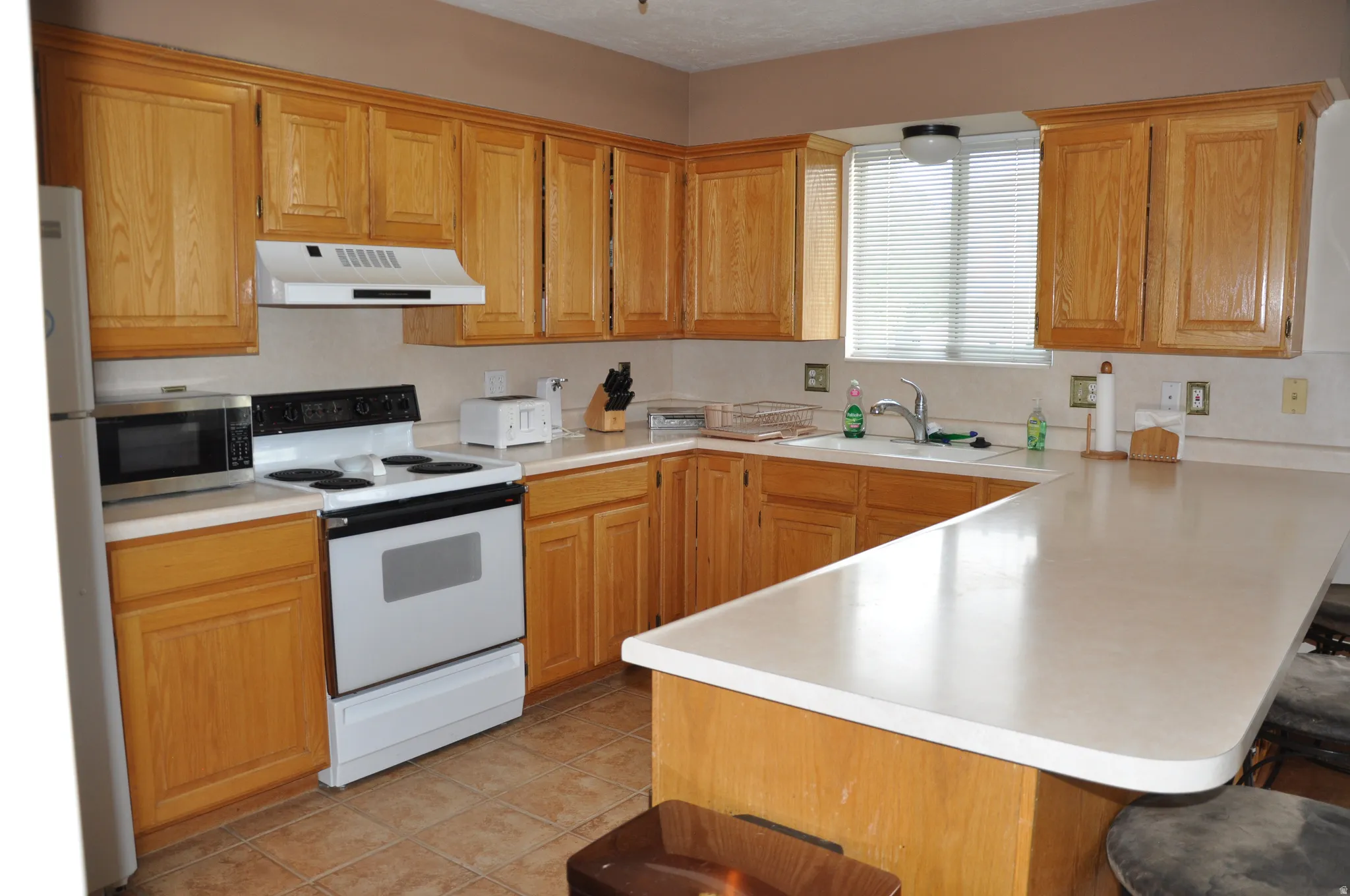 Spacious kitchen featuring extensive wood cabinetry, light-toned countertops, a white electric range, and a built-in breakfast bar