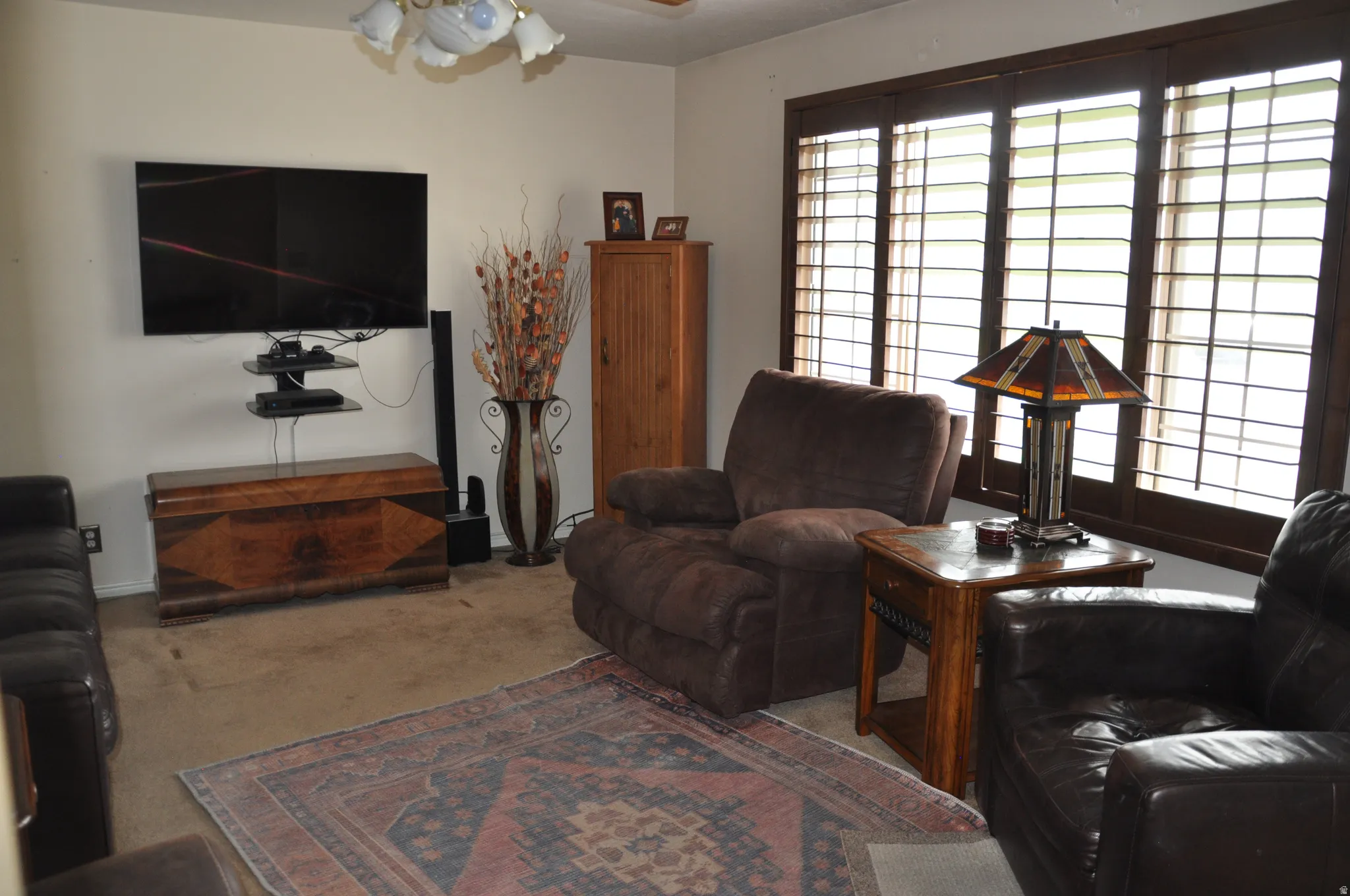 Wall-mounted television with shelving, wide windows with wood-tone plantation shutters, and a chandelier-style ceiling fixture