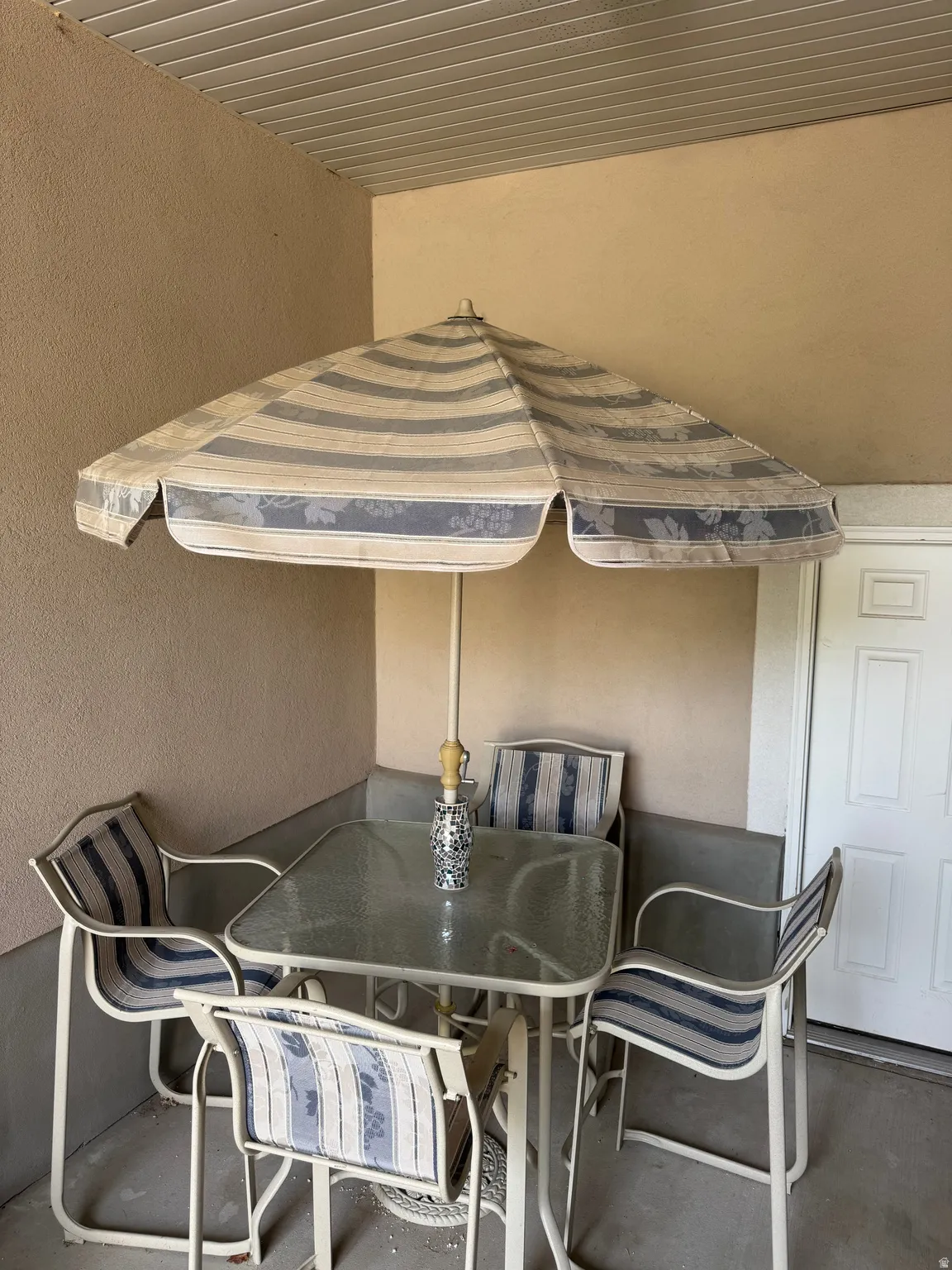 Covered outdoor dining area featuring a striped market umbrella, glass-top table, and aluminum frame seating for four