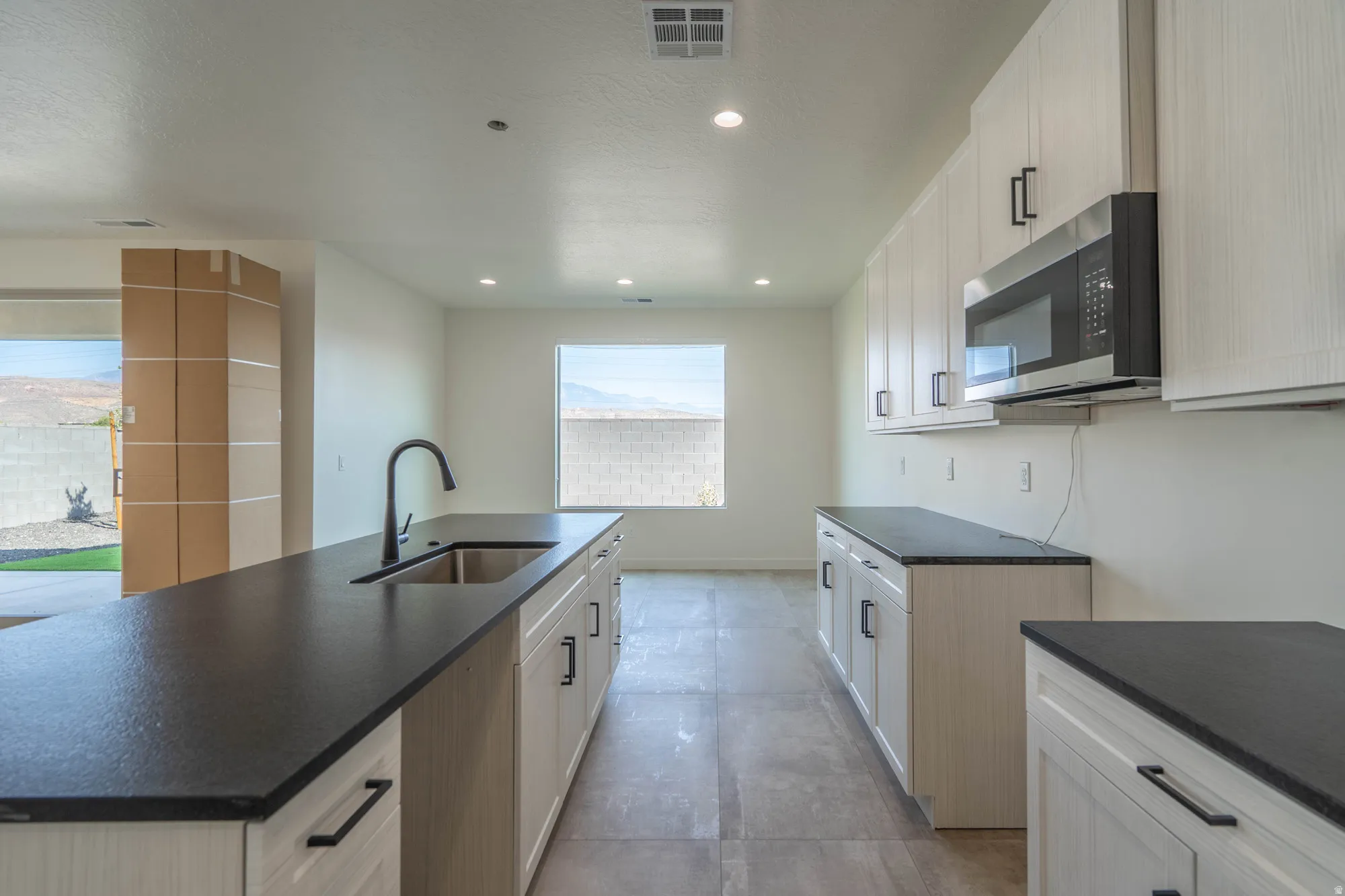 Kitchen featuring extensive light-toned cabinetry, dark countertops, a stainless steel microwave, a built-in sink with a gooseneck faucet, and recessed lighting