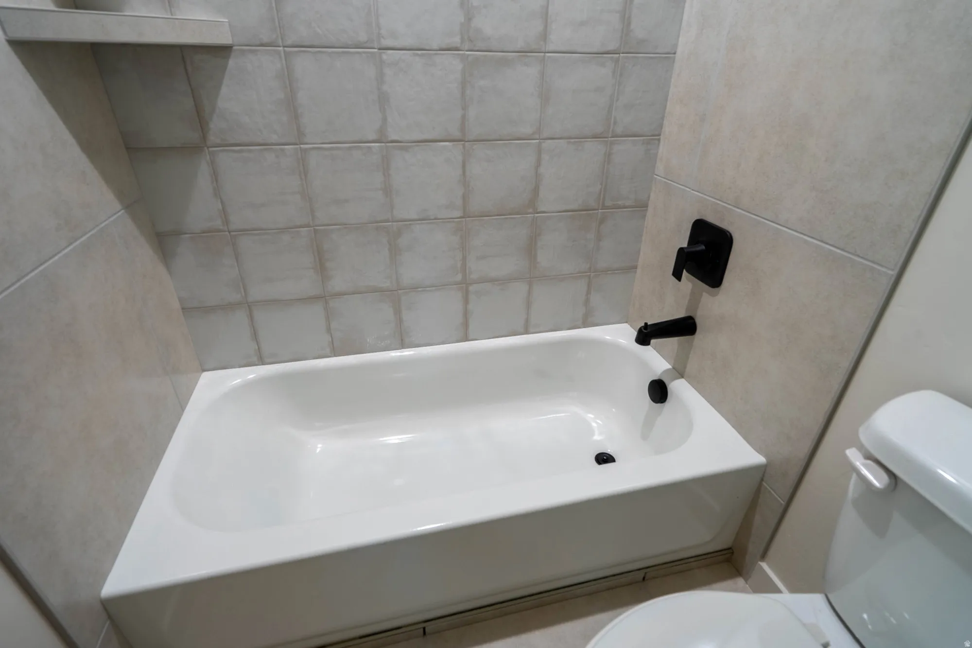 Bathtub with matte black fixtures, light-toned wall tiling, and a built-in shower shelf
