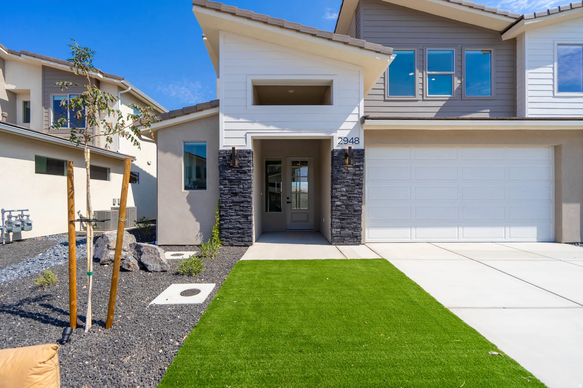 Contemporary exterior featuring a multi-tone facade, stone accents, a two-car garage, and a low-maintenance artificial turf lawn