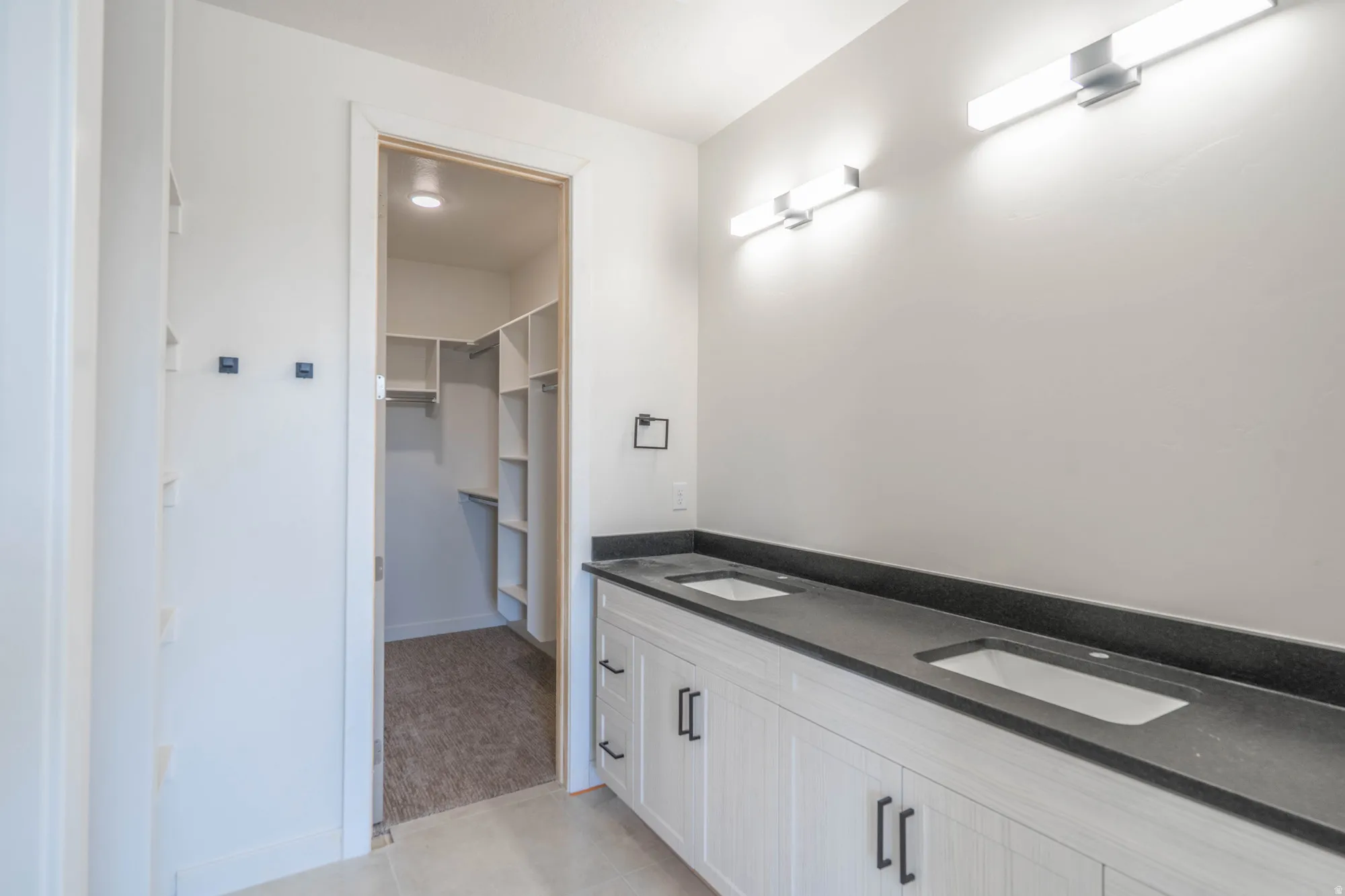 Dual vanity with dark countertops and integrated sinks, modern wall-mounted lighting, and an adjacent walk-in closet with built-in shelving