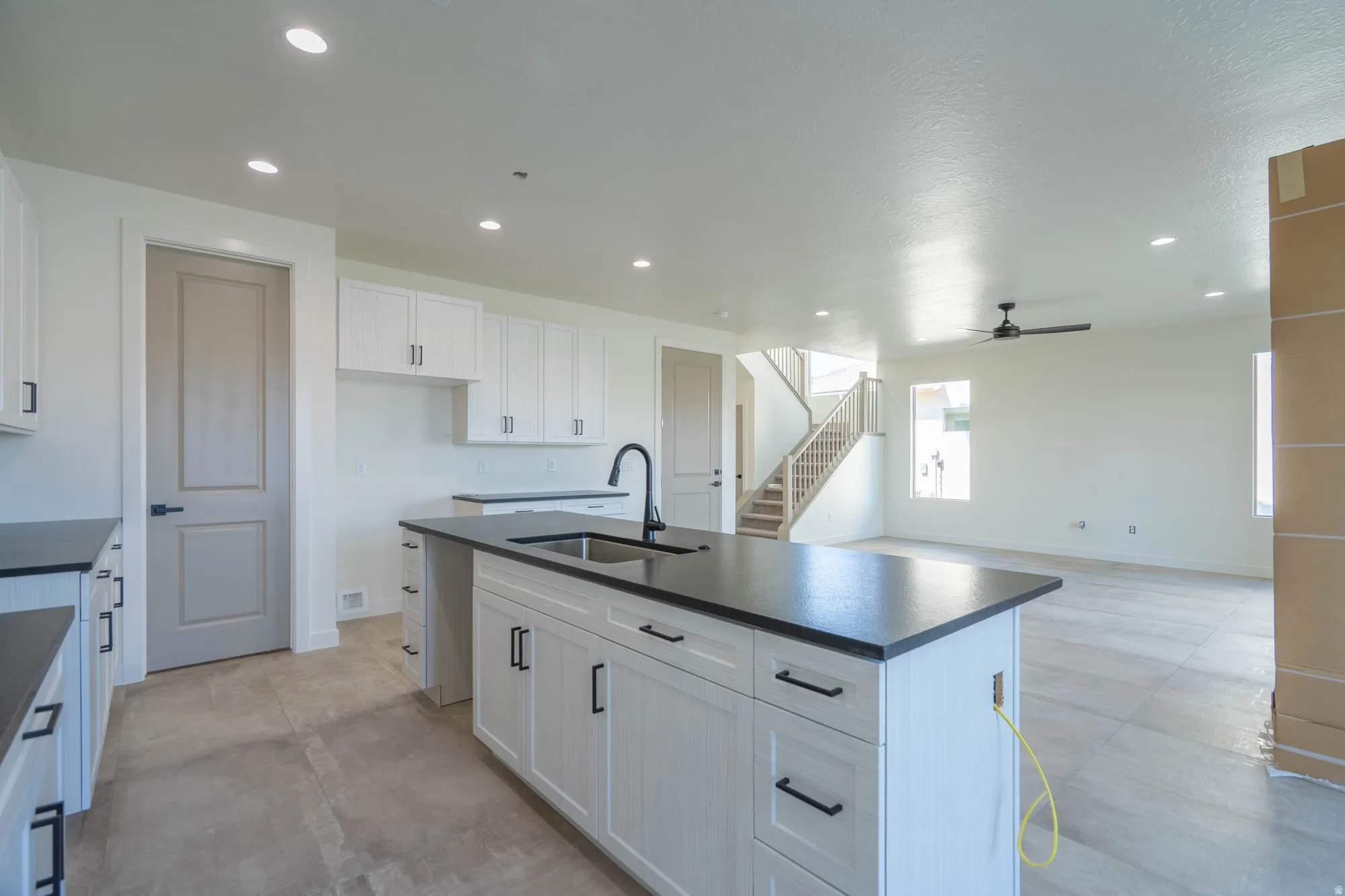 Chef's kitchen featuring a large center island with black quartz countertop and stainless steel sink, white shaker-style cabinetry with hardware accents, recessed ceiling lighting, and open sight lines to the great room and staircase beyond