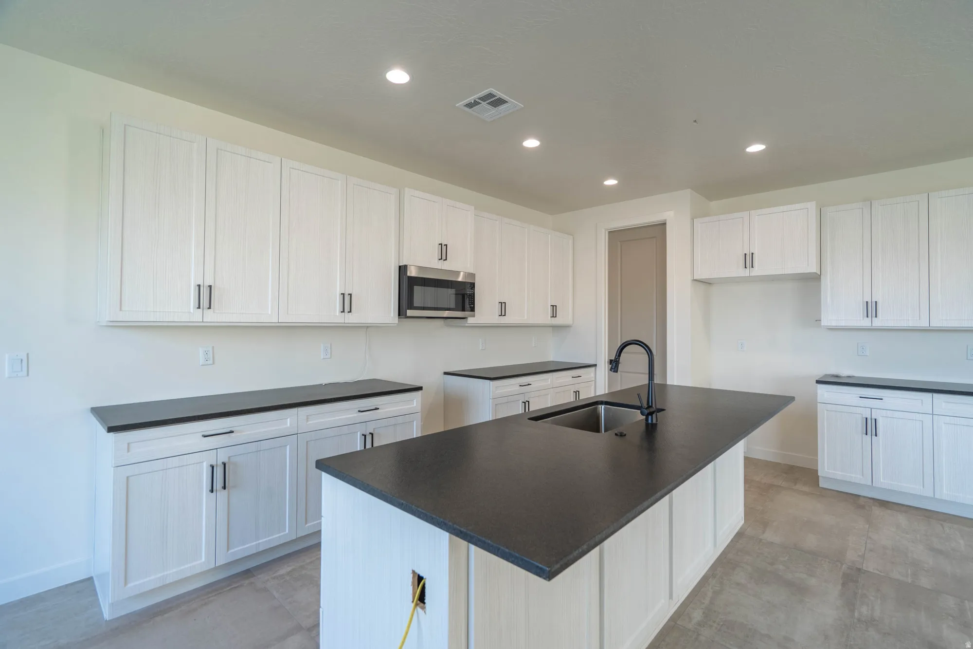 White cabinetry, dark countertops, kitchen island with integrated sink, stainless steel microwave, recessed lighting