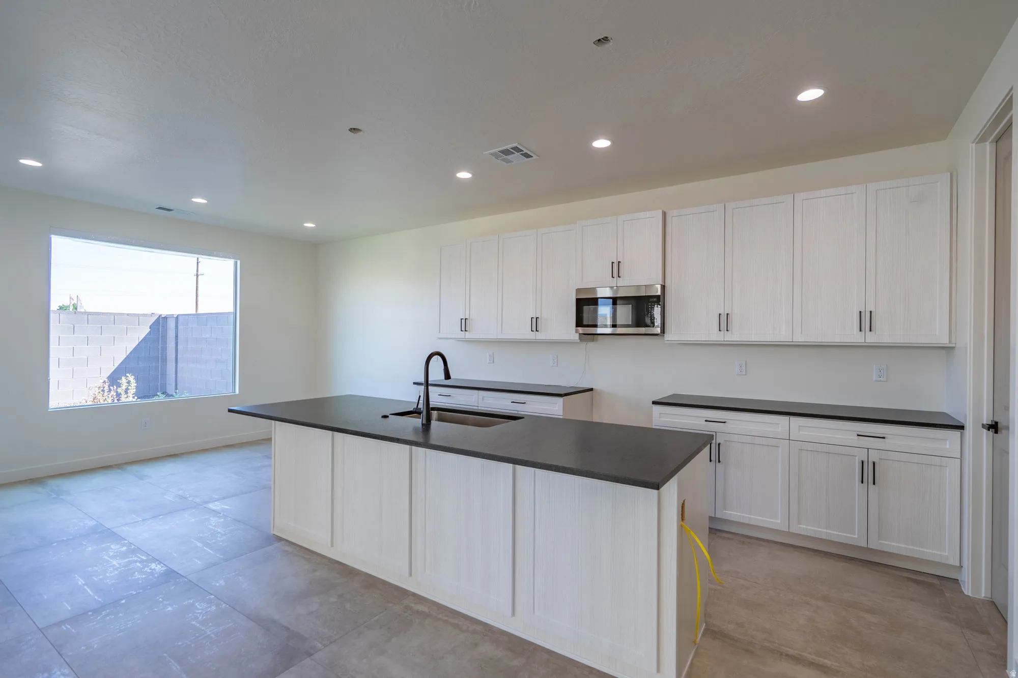 Kitchen featuring white shaker-style cabinetry, dark countertops, a stainless steel microwave, and an island with an integrated sink and matte black faucet