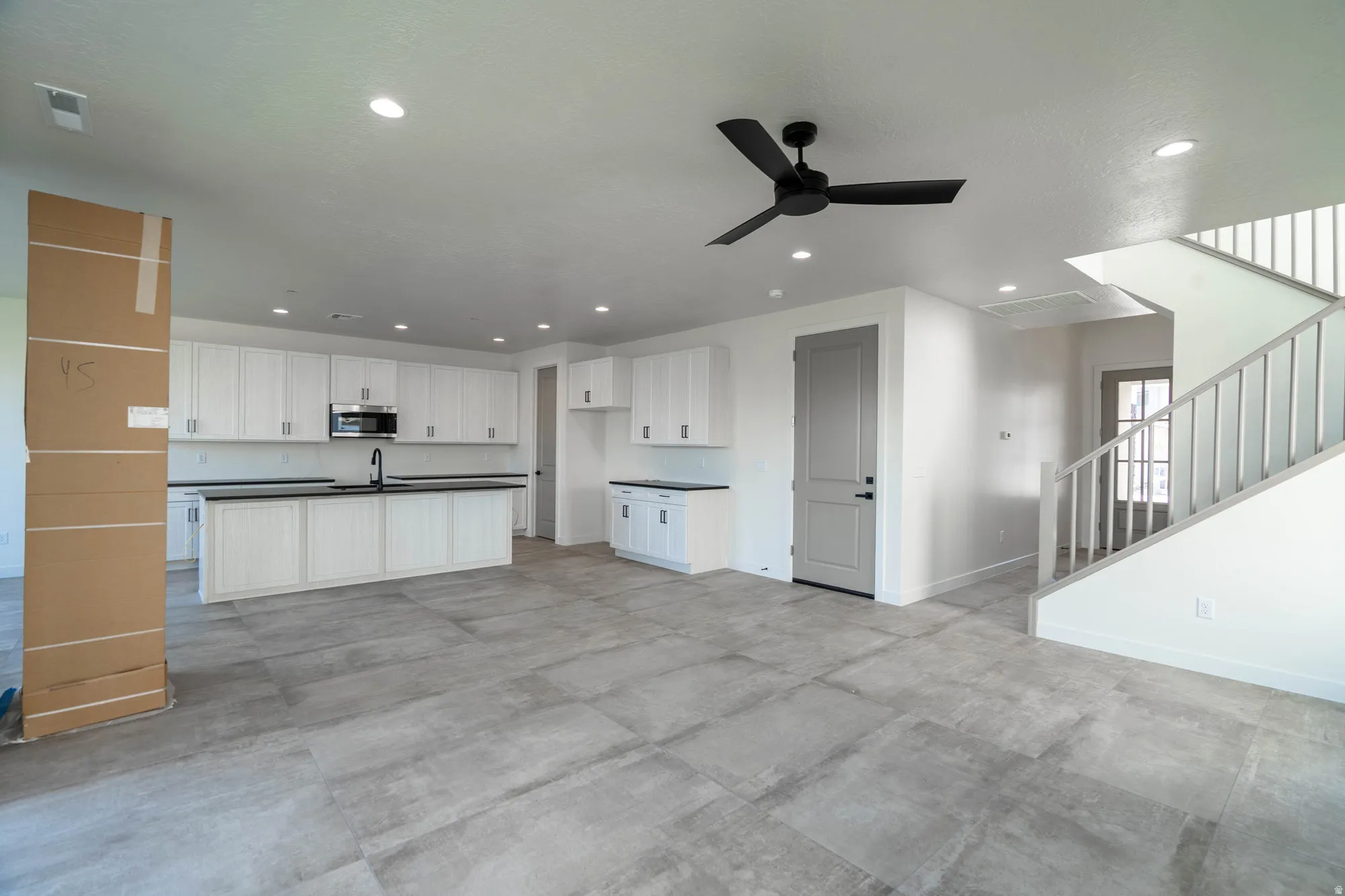 Spacious open-concept living area featuring large format tile flooring, a modern black ceiling fan, recessed lighting, and a kitchen with white cabinetry and dark countertops