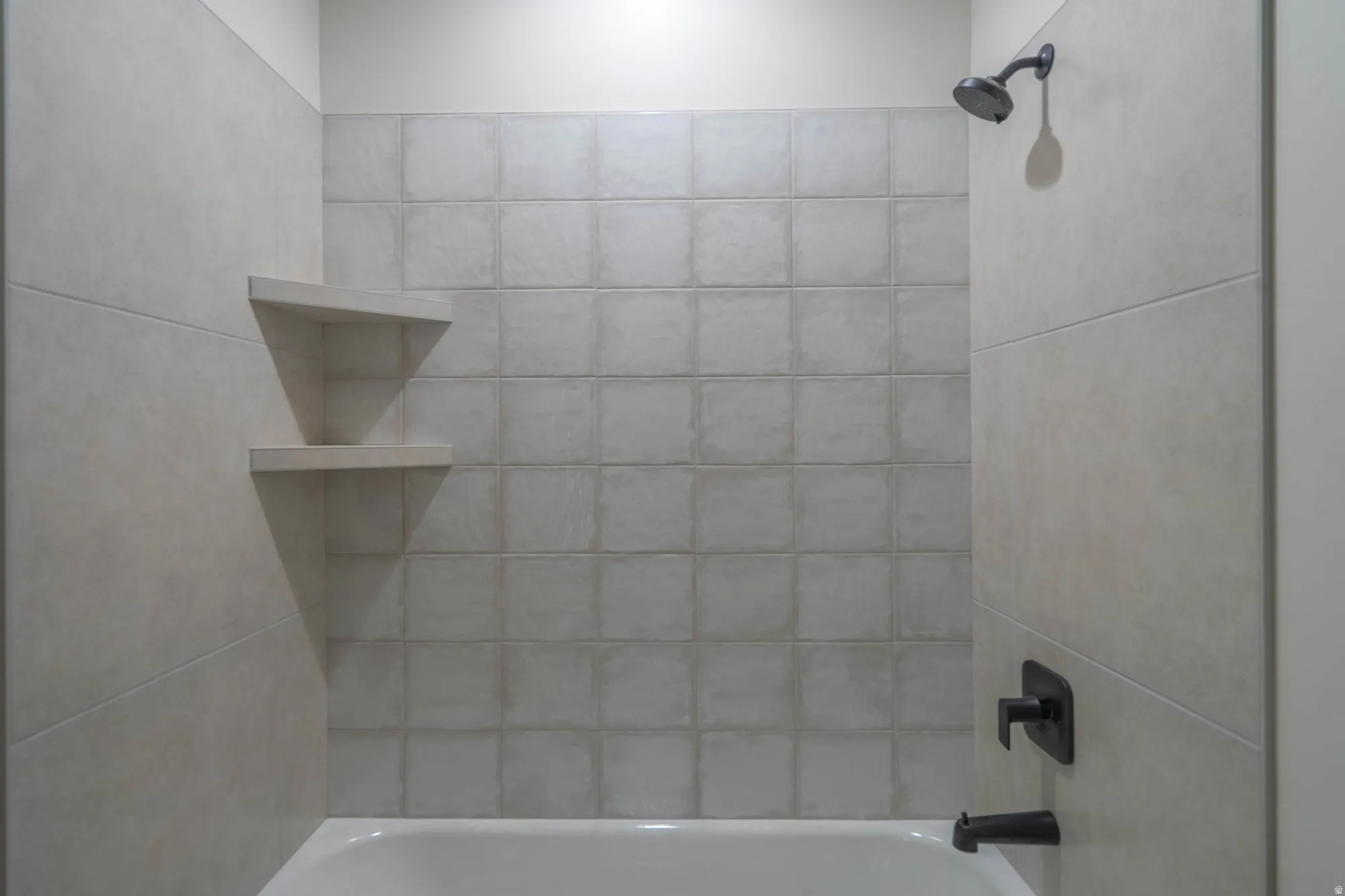 Bathtub with textured tile surround, dark-finish shower head, dark-finish faucet, and integrated corner shelving