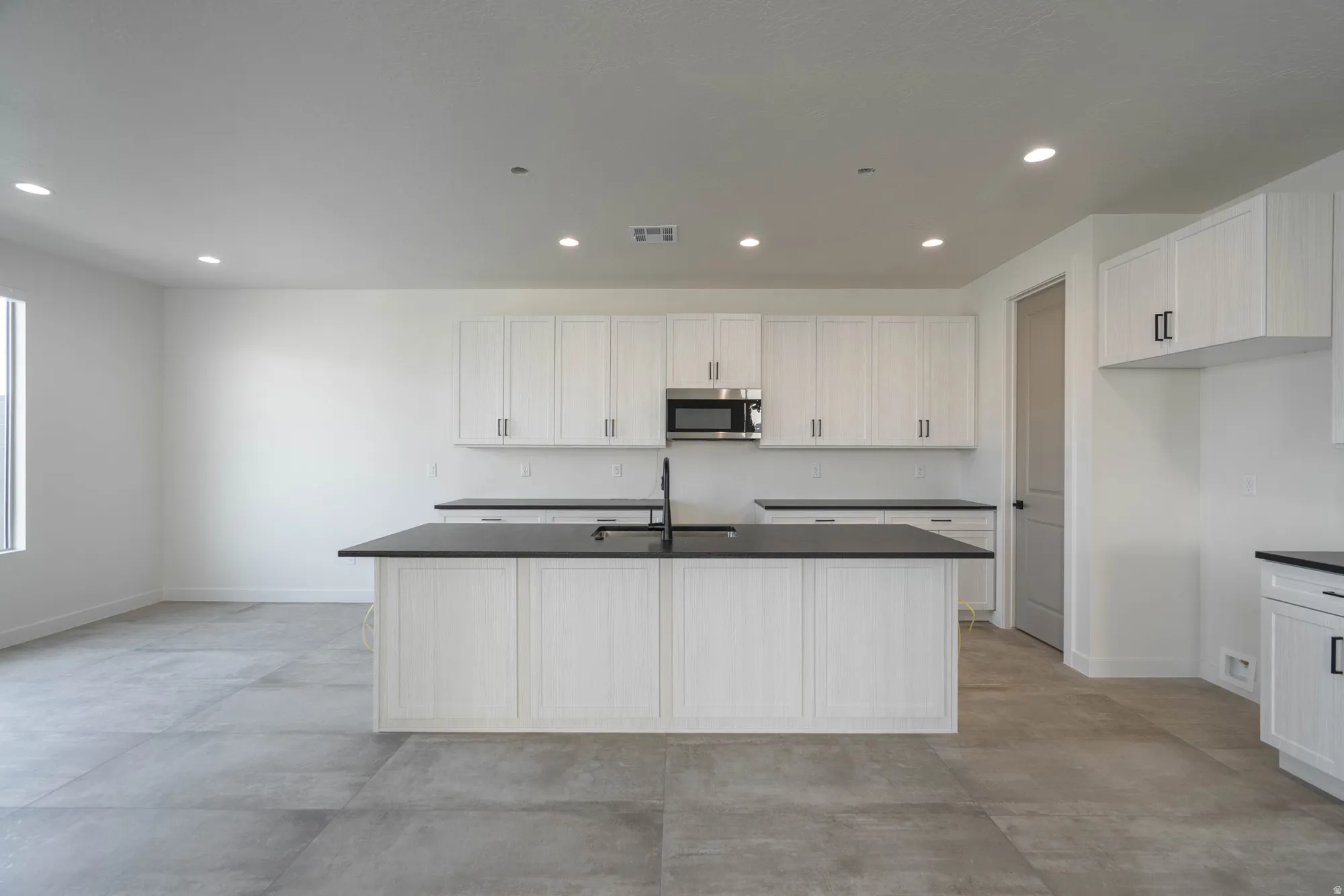 Kitchen featuring white cabinetry, dark countertops, a central island with an undermount sink, a built-in microwave, and large format tile flooring