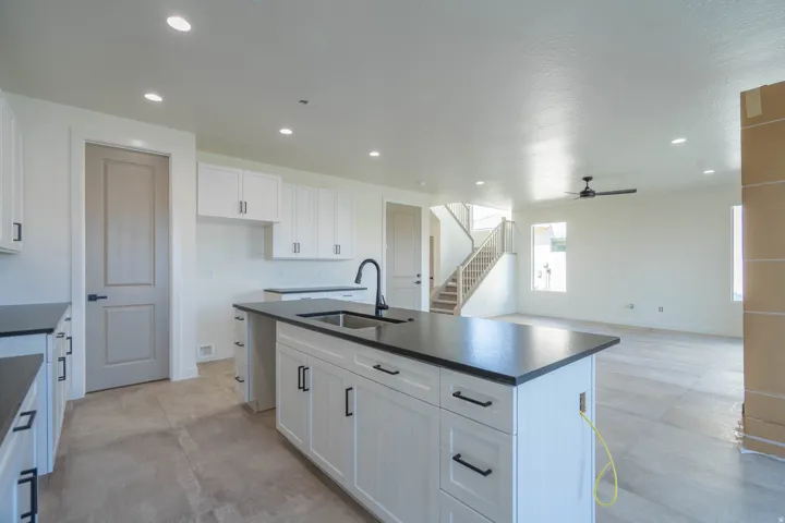 Chef's kitchen featuring a large center island with black quartz countertop and stainless steel sink, white shaker-style cabinetry with hardware accents, recessed ceiling lighting, and open sight lines to the great room and staircase beyond