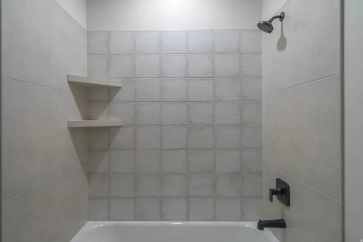 Bathtub with textured tile surround, dark-finish shower head, dark-finish faucet, and integrated corner shelving