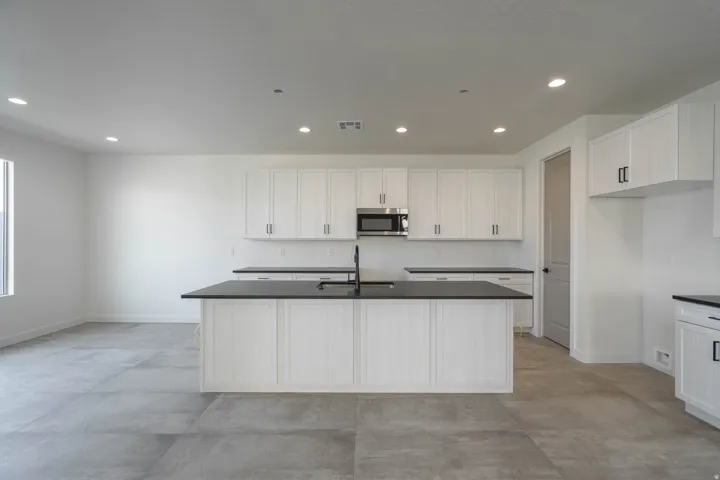Kitchen featuring white cabinetry, dark countertops, a central island with an undermount sink, a built-in microwave, and large format tile flooring