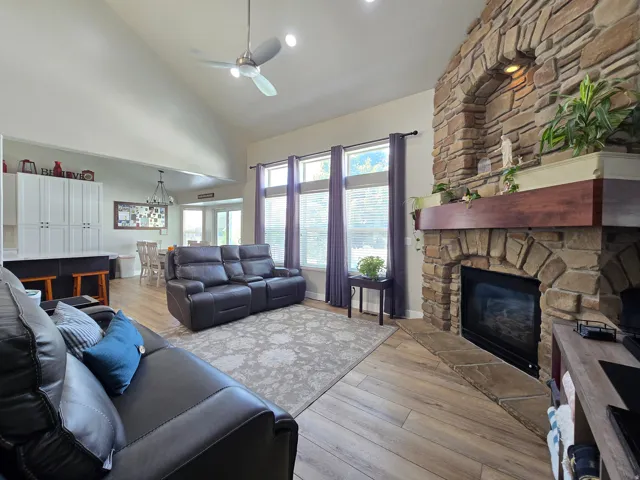 Vaulted ceiling with a ceiling fan, stone fireplace with a wood mantel, wood-finish flooring, and multiple windows