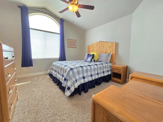 Bright secondary bedroom featuring an arched window with blue drapery, wood-finish nightstand, carpet flooring, and ceiling fan with integrated lighting
