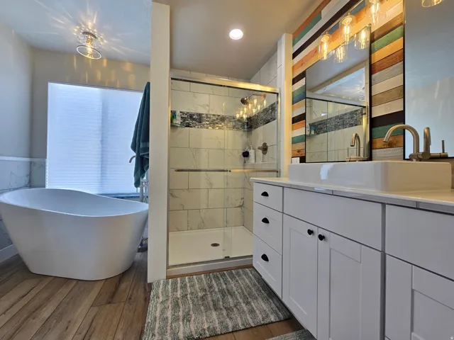 Freestanding soaking tub, walk-in shower with tile surround, wood-finish flooring, dual vanity with vessel sinks, and accent wall with wood paneling
