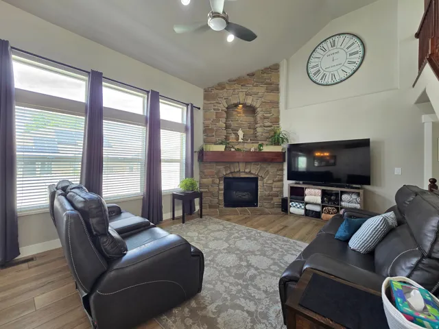 Vaulted ceiling with a ceiling fan, stone fireplace with a wood mantel, wood-finish flooring, and multiple windows