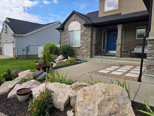 Stone veneer accents on the facade, arched window, covered entryway with columns, concrete walkway with paver inserts, and layered landscaping with large boulders