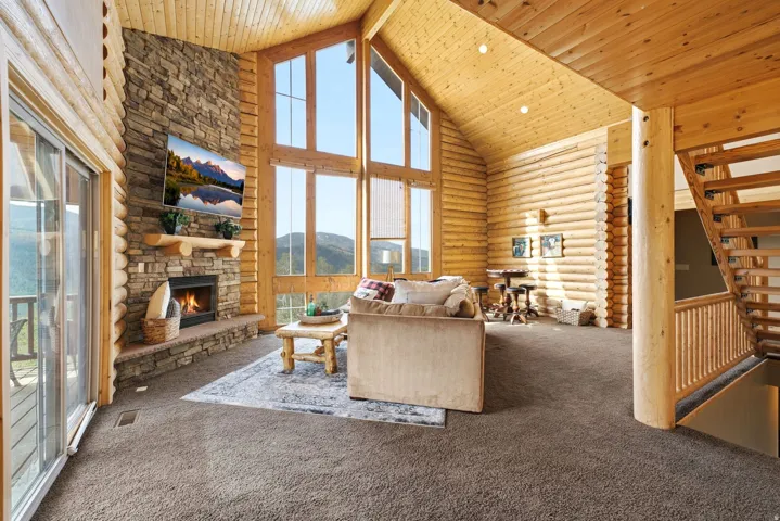 Vaulted ceilings, a stacked stone fireplace with a wood mantel, a wall of multi-pane windows, wood-paneled walls, and open-tread stairs with wood railings