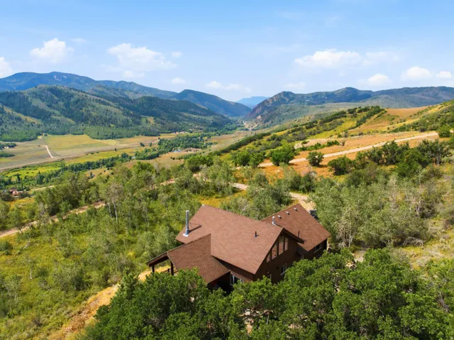 Terraced property with a brown-shingled roof and wood siding, surrounded by mature trees and natural landscaping, set against a backdrop of distant mountains