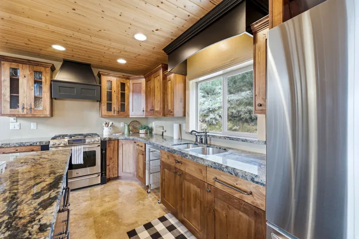 Kitchen featuring a wood-panel ceiling, stainless steel appliances, granite-style countertops, wood cabinetry with glass-front doors, and a double basin sink