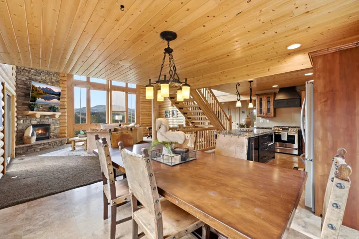 Open-concept living area with wood plank ceilings, a stone fireplace, large windows, a wood staircase, and a kitchen featuring a central island